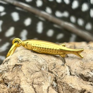 以下が含まれることがあります： 金色の魚のペンダント。ひれやうろこなど、細部までこだわったデザイン。小さなループから吊り下げられています。背景には羽と木の切れ端があります。