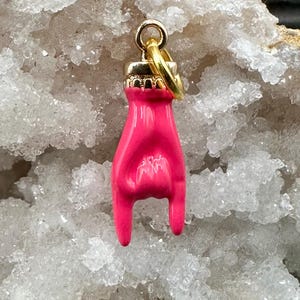 Puede incluir: Un dije rosa brillante con forma de mano haciendo el gesto de «Te amo». El dije tiene una parte superior dorada y un anillo dorado para sujetarlo. El fondo es una formación de cristales blancos y grises.