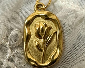 May include: A gold-toned pendant featuring a raised tulip design. The pendant is oval-shaped and has a textured surface.