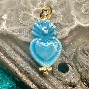 May include: A light blue ceramic pendant with a heart design. The pendant has a gold-colored bail and a decorative gold-colored bead at the bottom. The pendant is approximately 1.5 inches in length.