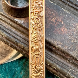 May include: Gold-toned pendant with the word "LEO" and a lion design. The pendant is intricately detailed with floral motifs.