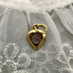 May include: A gold heart-shaped charm with a purple gemstone inside. The charm has a loop at the top for attaching to a necklace or bracelet.
