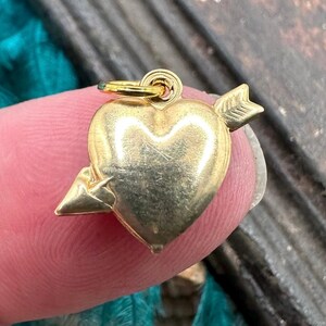 May include: A gold-coloured heart charm with an arrow piercing through it. The charm has a loop at the top for attaching to a necklace or bracelet.