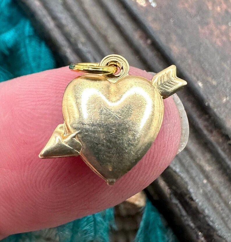 May include: A gold-coloured heart charm with an arrow piercing through it. The charm has a loop at the top for attaching to a necklace or bracelet.