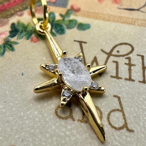 May include: A gold-colored starburst pendant with a central, oval-shaped, clear stone. The starburst has six points and is adorned with small, clear accent stones. The pendant is suspended from a small, gold-colored loop.