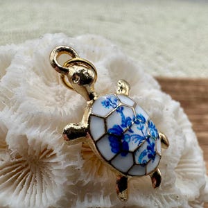 May include: A gold-tone turtle charm with a white and blue porcelain design. The turtle's shell is decorated with a floral pattern.