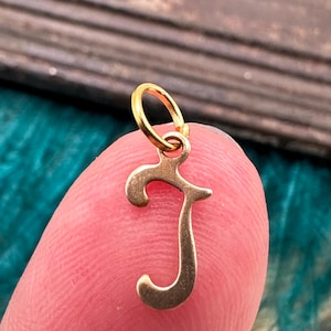 May include: Gold charm in the shape of the letter J. The charm has a loop at the top for attaching to a necklace or bracelet.