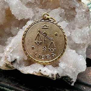 May include: Gold-toned pendant featuring the astrological symbol for Libra, a set of scales, with the word "LIBRA" inscribed below. The pendant is on a white, crystalline background.