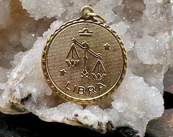 May include: Gold-toned pendant featuring the astrological symbol for Libra, a set of scales, with the word "LIBRA" inscribed below. The pendant is on a white, crystalline background.