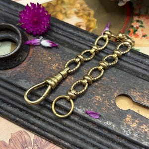 May include: Two antique brass chain clasps with a twisted rope design and a spring clasp and ring. The clasps are on a dark, textured surface with a purple flower and petals.
