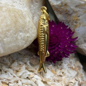 May include: A gold-colored fish pendant with detailed scales and fins. The pendant is suspended from a small loop at the top. The background includes a purple flower and a light-colored stone.