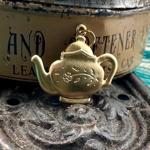 Puede incluir: Un amuleto de tetera de color dorado con un diseño floral. El amuleto cuelga de una cadena y descansa sobre una superficie oscura.