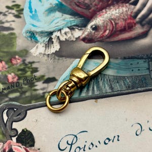Puede incluir: Un mosquetón giratorio dorado con una puerta con resorte y un pequeño anillo. El mosquetón es de metal y tiene un acabado brillante. El fondo es una postal vintage con una imagen y texto descoloridos.
