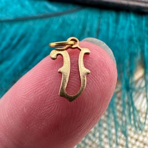 May include: Gold charm in the shape of the letter "U". The charm is ornate and has a loop at the top for attaching to a necklace or bracelet.