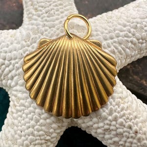 Puede incluir: Un colgante dorado con forma de concha, con un pequeño lazo para una cadena. El colgante tiene un diseño detallado y acanalado, sobre un fondo de estrella de mar blanca. Es una joya.