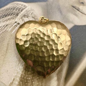 May include: A large, gold-colored heart-shaped pendant with a hammered texture. The pendant has a loop at the top for attaching to a chain.