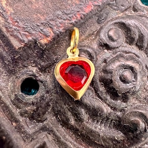 Puede incluir: Un colgante de corazón de oro con una gema de vidrio roja en el centro.