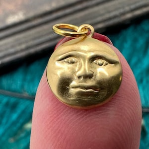 May include: A gold-colored pendant charm in the shape of a face with a round, moon-like outline. The face has a serious expression with closed lips and slightly raised eyebrows.