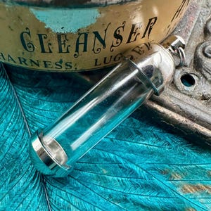 May include: A silver and glass vial pendant with a clear glass tube and a silver cap. The pendant is on a blue and green background.