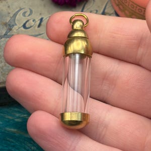 May include: A small, clear glass vial with gold-coloured metal caps and a loop at the top. The vial is cylindrical and appears empty. The gold caps are rounded and smooth. The vial is held in a person's hand.