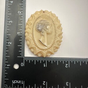 May include: A cream-colored cameo brooch with a raised design of a woman's profile. The woman is wearing a crown and has a beaded necklace. The cameo is surrounded by a decorative border with gold accents.