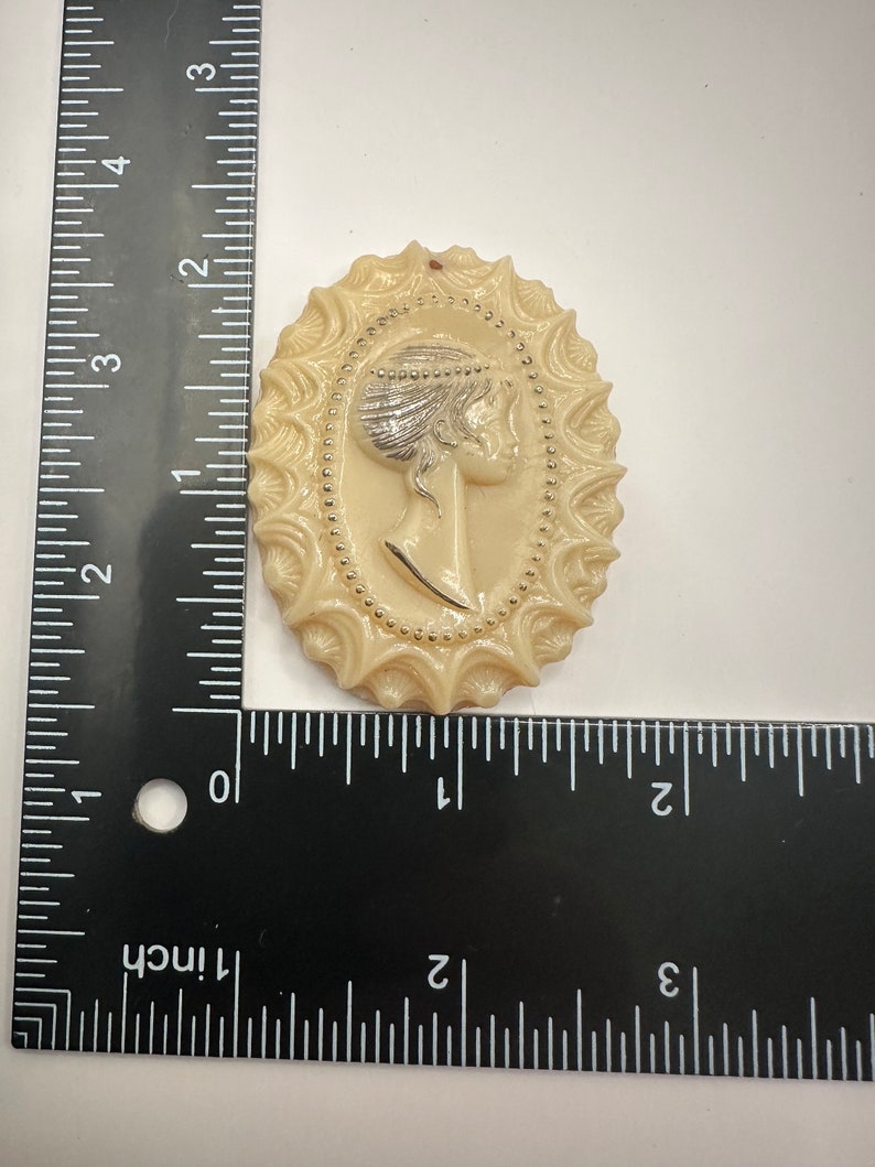 May include: A cream-colored cameo brooch with a raised design of a woman's profile. The woman is wearing a crown and has a beaded necklace. The cameo is surrounded by a decorative border with gold accents.