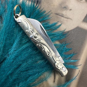 May include: A silver pocket knife with a floral design and a gold ring attached to the top. The knife is lying on a bed of teal feathers.