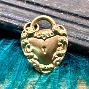 Puede incluir: Un dije de candado en forma de corazón de oro con detalles intrincados y una cerradura. El dije está sobre un fondo de plumas color turquesa.