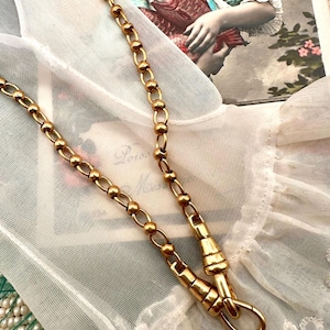 May include: A gold-coloured necklace featuring a chain of alternating oval and round links. The necklace has a clasp and is displayed on white, sheer fabric. A vintage photograph is visible in the background.