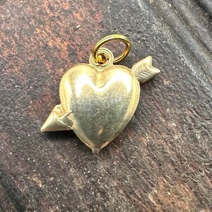 May include: A gold heart charm with an arrow through it. The charm has a loop at the top for attaching to a necklace or bracelet.