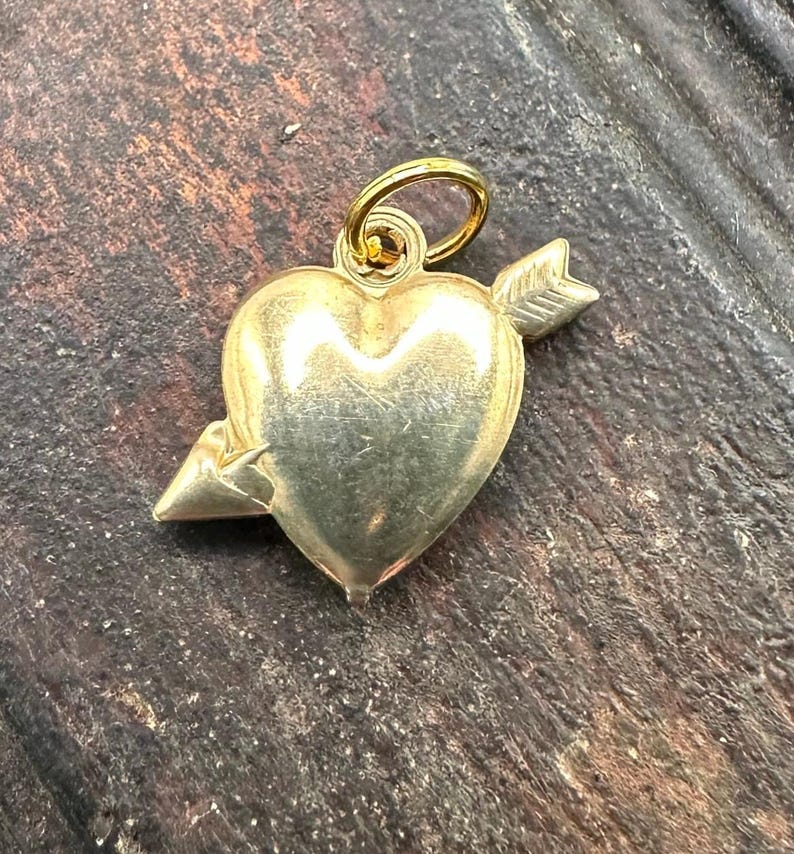 May include: A gold heart charm with an arrow through it. The charm has a loop at the top for attaching to a necklace or bracelet.