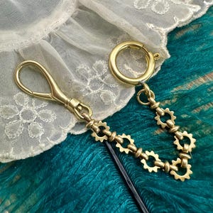 May include: A brass-colored keychain with a lobster clasp, a decorative chain of gear-shaped links, and a large circular clasp. The keychain rests on a teal surface with a white lace trim.