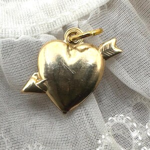 May include: A gold heart charm with an arrow through it. The charm is on a white background.