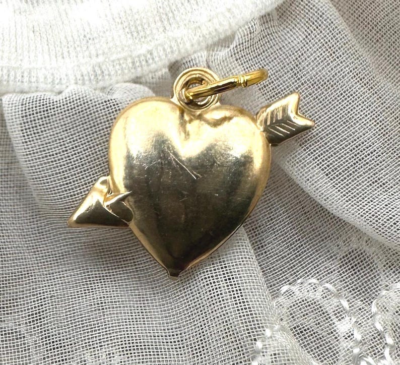 May include: A gold heart charm with an arrow through it. The charm is on a white background.