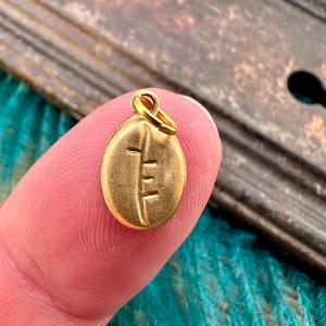 May include: A gold-coloured coffee bean charm with a detailed design. The charm has a loop for attaching to a necklace or bracelet.