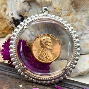 May include: A circular pendant featuring a 1975 United States copper penny encased in a clear, domed glass setting. The coin is surrounded by a silver-toned metal frame with a beaded edge and a small jump ring at the top for attachment.