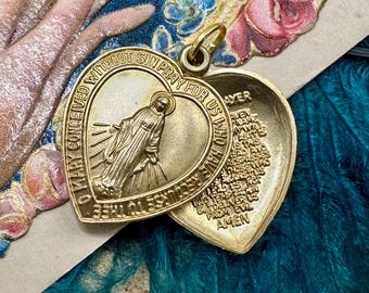 May include: A gold-colored heart-shaped locket with an image of a religious figure and text. The locket is open, revealing an inscription inside. The text around the edge reads "O Mary conceived without sin pray for us who have recourse to thee."