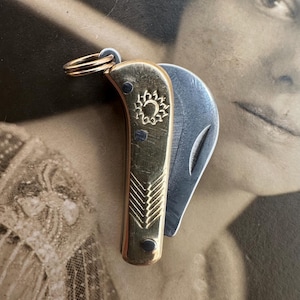 Puede incluir: Un pequeño cuchillo de bolsillo antiguo de latón con una hoja de plata. El cuchillo tiene un diseño decorativo en el mango y un anillo para sujetarlo a un llavero.