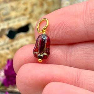 May include: A small bell-shaped charm featuring a deep red glass bead with gold-colored metallic accents and a gold-colored jump ring attached to the top. The charm has a smooth, glossy finish and a small gold-colored bead at the bottom tip.