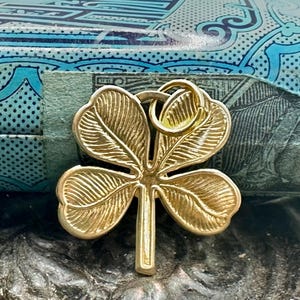 May include: A gold-colored four-leaf clover pendant with detailed veining on each leaf. The pendant has a small loop at the top for attaching to a chain or other jewelry. The clover is set against a backdrop of blue and green.