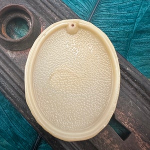 May include: A white oval ceramic incense burner with a small hole in the center. The burner has a textured surface and is sitting on a wooden surface.