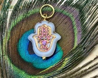 May include: A translucent white Hamsa hand charm featuring a gold-toned metal outline and a central eye motif with iridescent purple and gold detailing. The charm is attached to a circular gold-toned jump ring at the top for easy attachment.