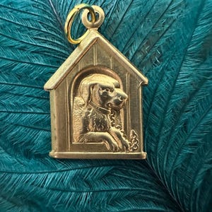 May include: Gold-toned charm of a dog inside a doghouse. The charm is detailed and has a 3D effect.