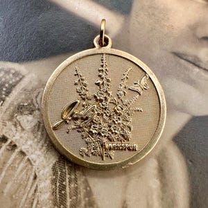 May include: Gold-toned pendant featuring a detailed floral design with the word "larkspur" inscribed at the bottom. The pendant is round and has a textured surface.