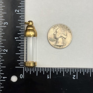 May include: A small, clear glass vial with a gold-coloured metal cap and base. The vial is approximately 5 cm tall. A US quarter coin is next to the vial for size comparison.