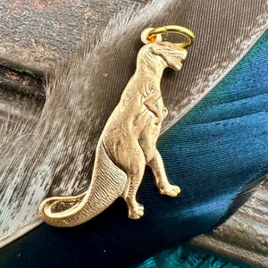 May include: Gold-colored dinosaur charm with a ring for attaching to a necklace or bracelet. The dinosaur is standing on all fours with its tail curled behind it.