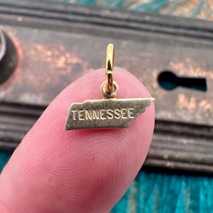 May include: A gold-colored charm in the shape of the state of Tennessee. The word "TENNESSEE" is engraved on the charm. The charm is suspended from a small loop, ready to be added to a necklace or bracelet.