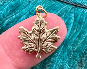 May include: A gold-colored maple leaf charm pendant. The leaf is detailed with a textured surface and has a loop at the top for attaching to a chain or cord.