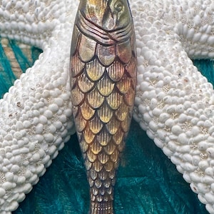 May include: A bronze and gold fish pendant with a loop for hanging. The pendant has a detailed scale pattern and is displayed on a white starfish and teal background. The fish is bronze, gold, and silver in colour.
