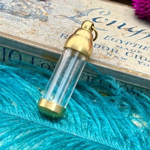 May include: A clear glass vial with brass-coloured metal caps and a loop at the top. The vial is cylindrical and sits on a bed of turquoise feathers. The background includes a vintage box with text.